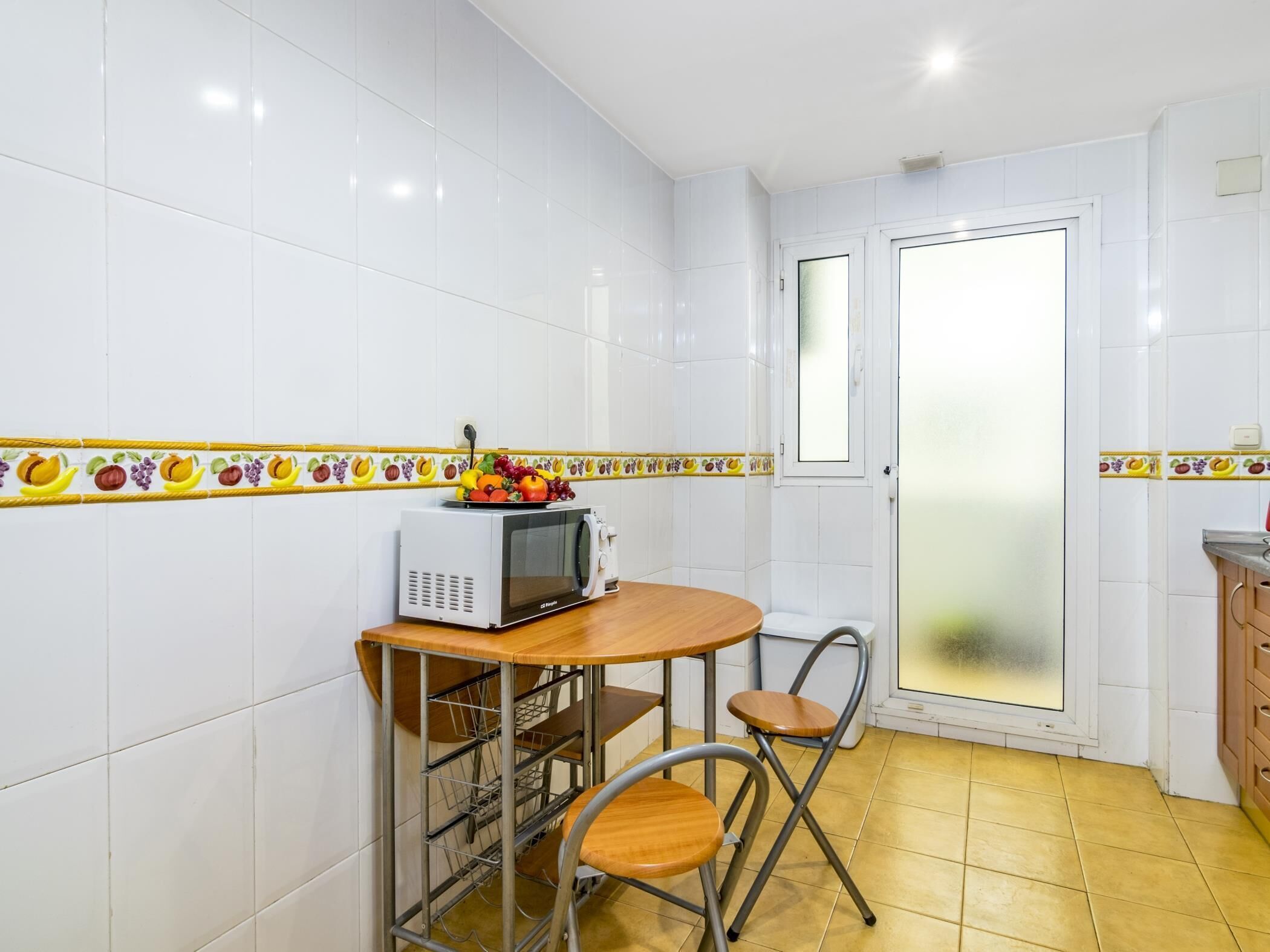 A compact kitchen with white tiled walls, a microwave on a wooden table, and a fruit bowl.