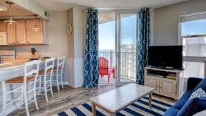A compact living space with a kitchen area, a dining table with chairs, a sofa, a TV, and a balcony with red chairs. - Adorable Updated Oceanfront Condo on the top floor! (North Topsail Beach)