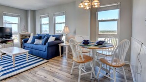 A living room with a blue sofa, white chairs, a dining table, and a TV. - Adorable Updated Oceanfront Condo on the top floor! (North Topsail Beach)