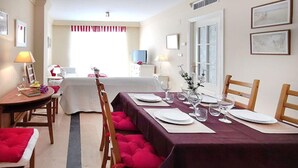 Uma sala de jantar com uma mesa comprida posta para refeição, cadeiras de madeira e uma janela com cortinas.