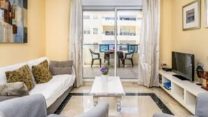 Uma sala de estar com sofá, mesa de centro, televisão e varanda com vista para edifícios.