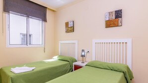 Quarto com duas camas individuais, cada uma com colcha verde e toalhas brancas. Há uma janela com persiana, uma mesa de cabeceira com abajur e duas pinturas penduradas na parede.