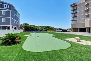 A mini-golf course with a putting green, surrounded by a white fence, and adjacent to a multi-story building with balconies.