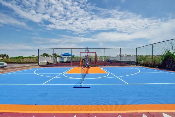 Un terrain de basket-ball en plein air, doté d’une surface de jeu bleue et orange, entouré d’une clôture en treillis métallique.
