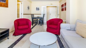 A living room with a white sofa, two red armchairs, a round coffee table, and a dining area with a table and chairs. - Banana Beach (Marbella)