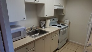 Een compacte keuken met witte kastjes, een magnetron, een spoelbak, een kookplaat en een plafondlamp.