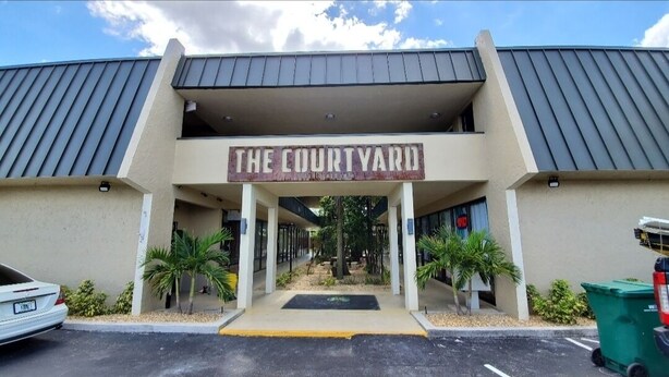Het uiterlijk van hotel The Courtyard, met een centrale ingang, palmbomen en een parkeerplaats.