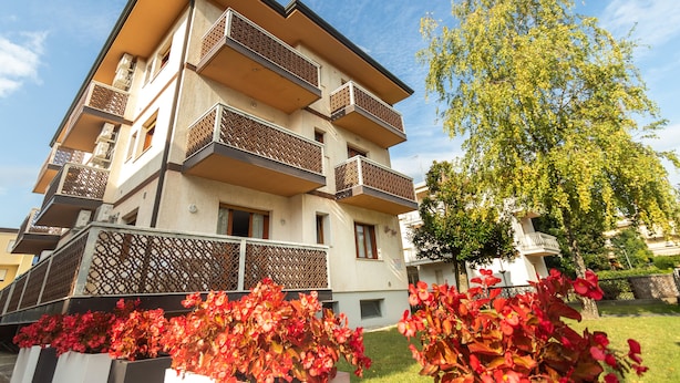 Un edificio residencial de varios pisos con balcones, un árbol y plantas con flores rojas en primer plano.