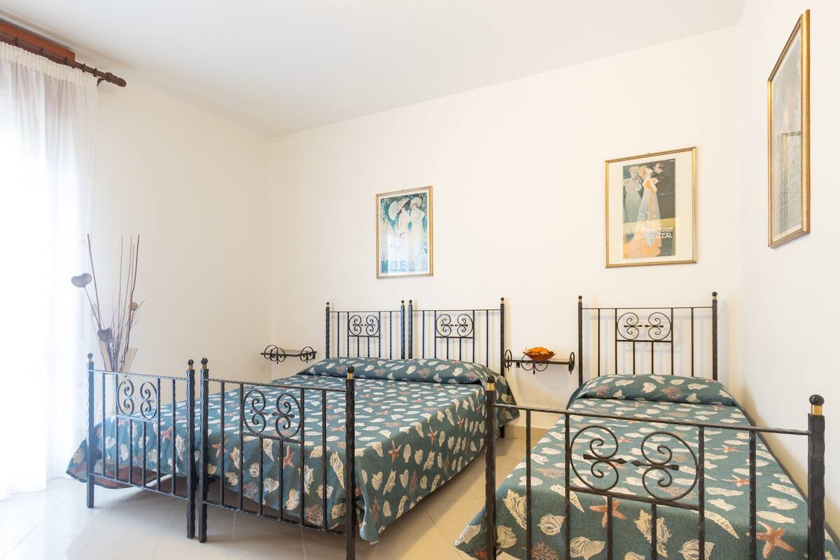 A bedroom with two beds, each with a metal frame and floral bedding. There are two framed pictures on the wall, and a window with a white curtain.