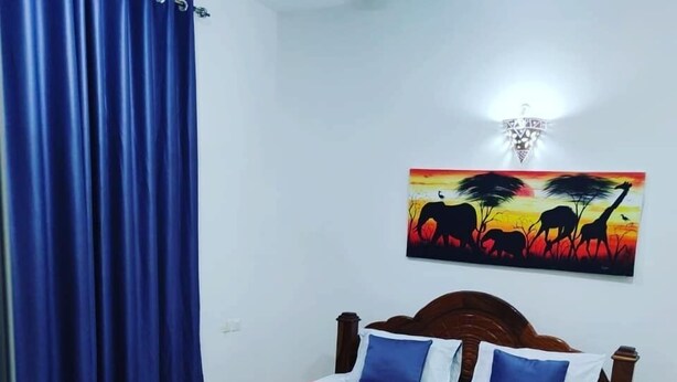 A neatly made bed with a blue and white bedspread, a wooden headboard, and a painting of a savannah scene on the wall. - Tropicana Kendwa Beach Hotel (Kendwa)