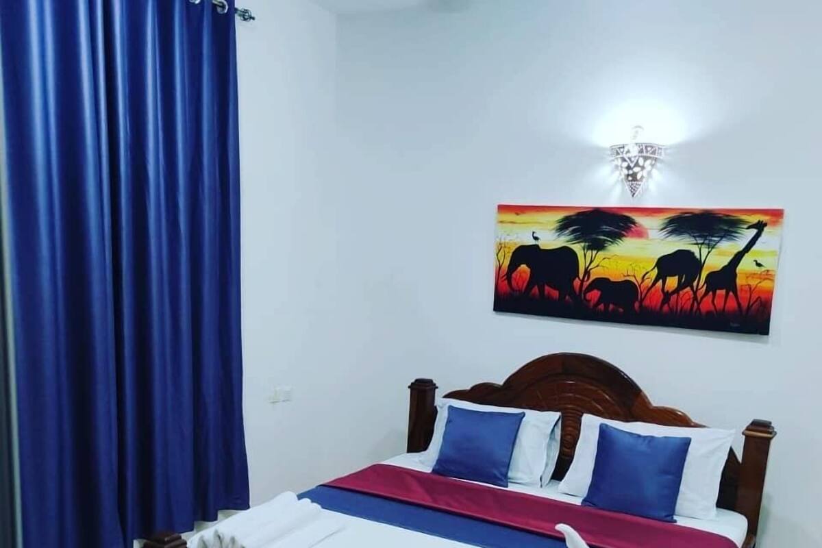 Ein ordentlich bezogenes Bett mit einer blau-weißen Tagesdecke, einem Holzkopfende und einem Bild einer Savannenlandschaft an der Wand.