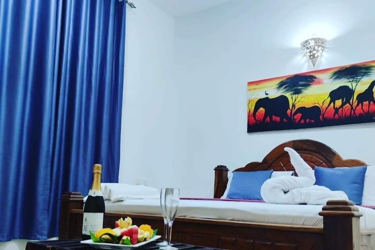 Ein Hotelzimmer mit einem Bett, einem Nachttisch mit Flasche und Gläsern, einem Elefantenbild und einem blauen Vorhang.