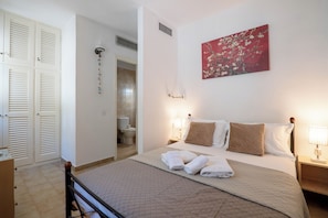 Habitación de hotel con cama, lámparas de noche, un cuadro, baño visible a través de una puerta abierta y un armario con puertas persianas.