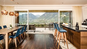 A modern dining area with a wooden table and chairs, a kitchen with a bar, and a scenic mountain view through large windows. - Chalet Penthouse   – Schenna Chalet (Schenna)
