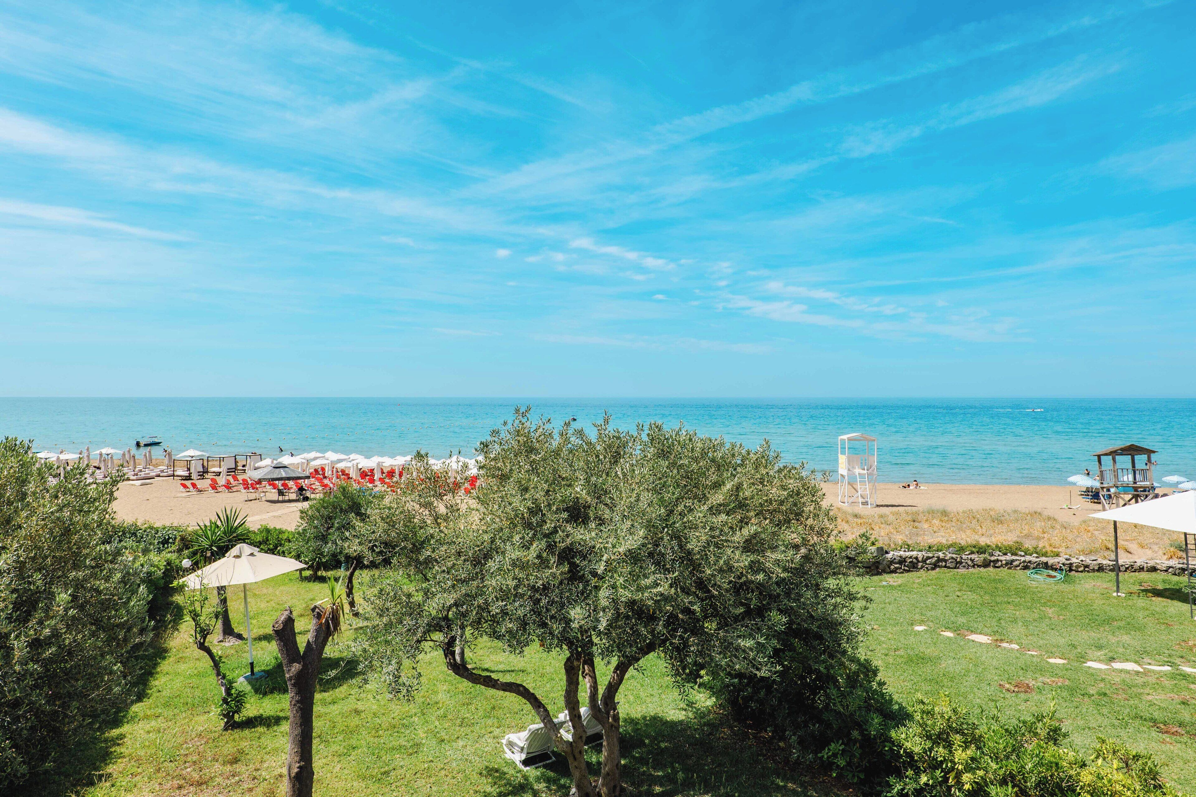 Vista de playa con zona verde, árboles y sombrillas.