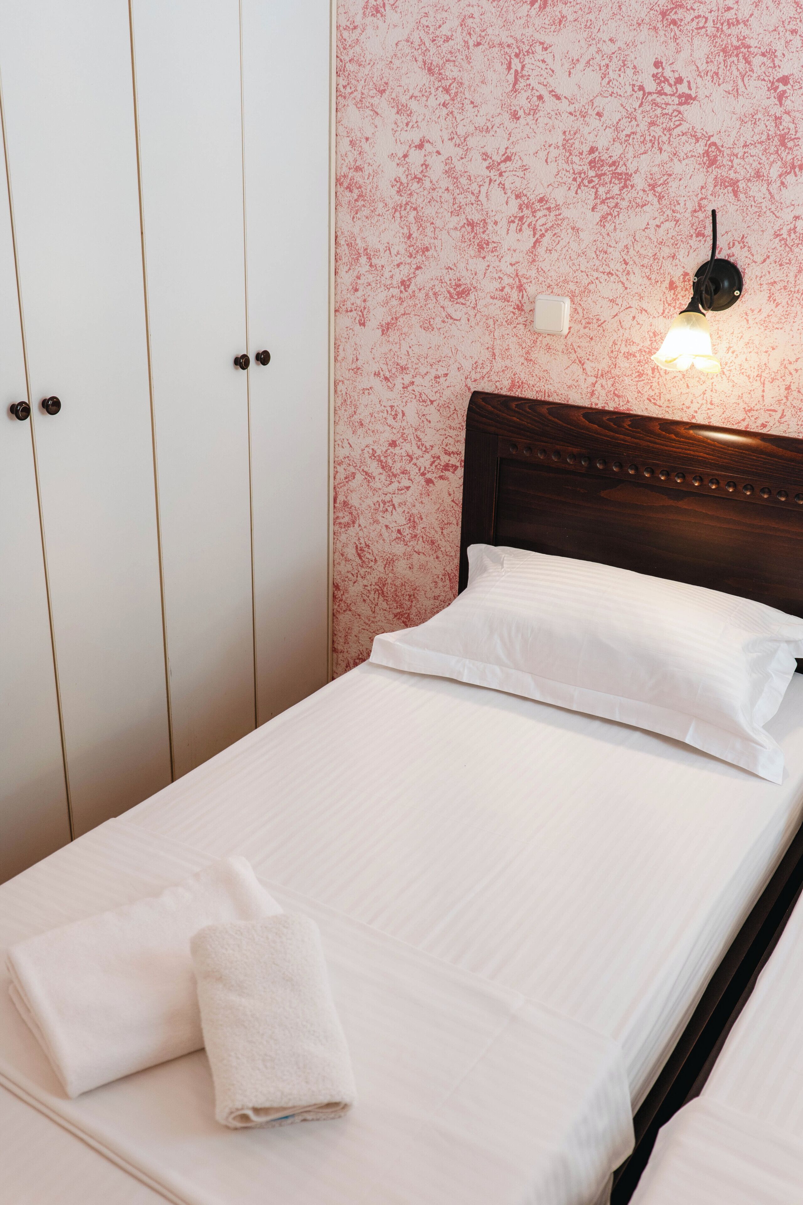 A neatly made bed with white linens and two towels, a wooden headboard, a wall-mounted light fixture, and a white wardrobe with a floral pattern.