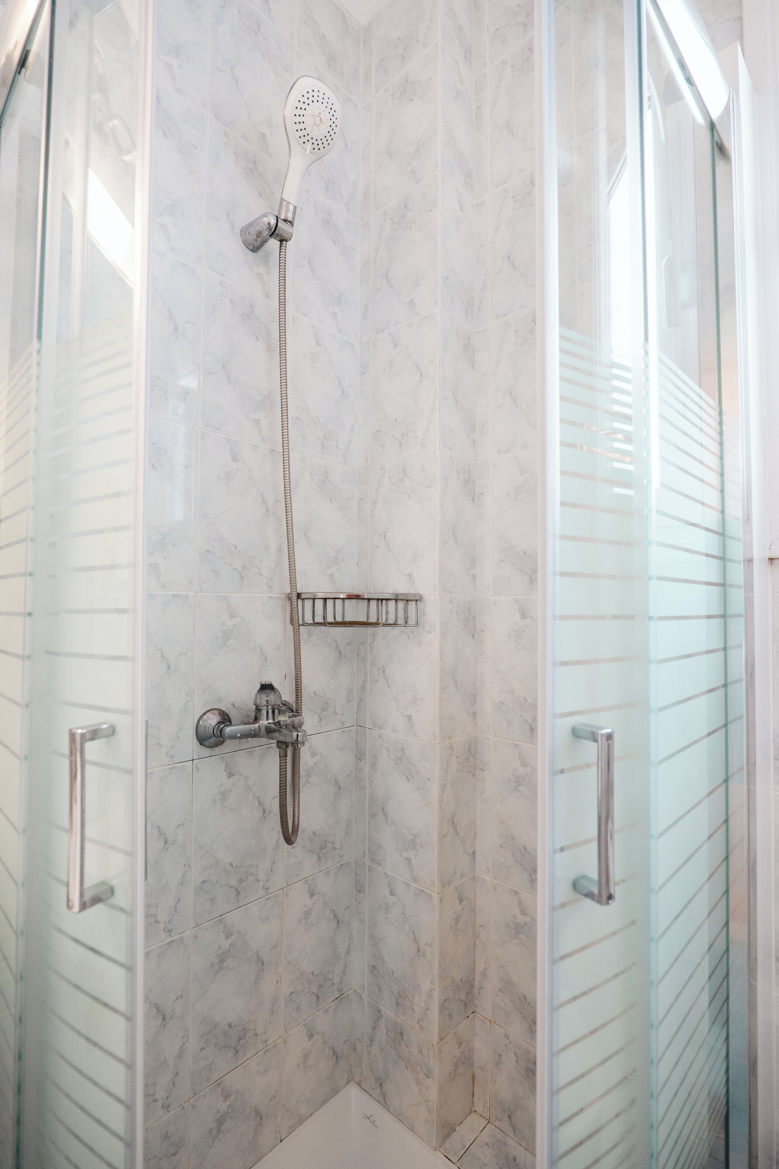 A glass-enclosed shower stall with a handheld showerhead, a wall-mounted faucet, and a small shelf.