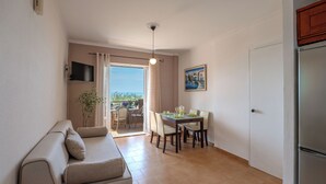 A compact living space with a sofa, dining table, chairs, a refrigerator, and a sliding glass door leading to an outdoor area. - Corfu Glyfada Menigos Resort 5-8-a (Corfu)