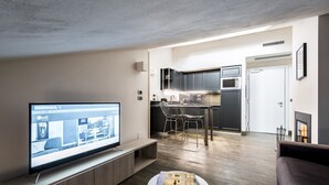 Una sala de estar moderna con un televisor de pantalla plana, una mesita redonda con una revista, un sofá, una zona de cocina con armarios oscuros y un comedor con sillas de metal.