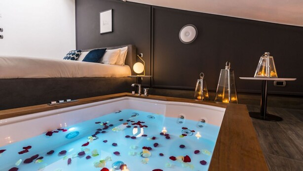 Una habitación con una bañera llena de agua y pétalos de rosa, una cama con cabecera oscura y una pequeña mesa con velas.