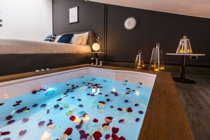 A room with a bathtub filled with water and rose petals, a bed with a dark headboard, and a small table with candles.