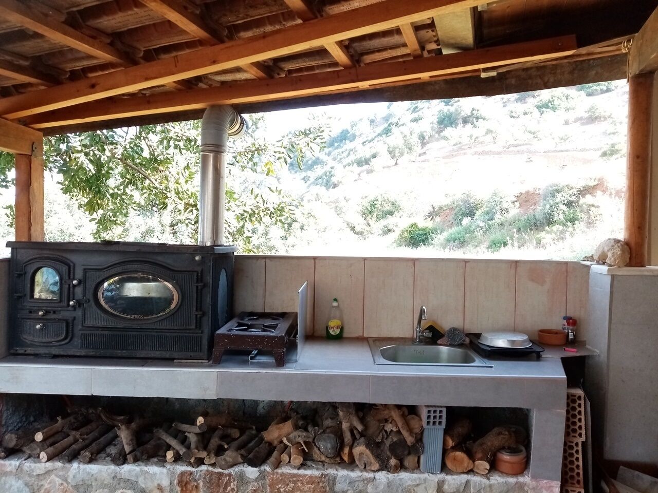 Cocina exterior con estufa de leña, pileta y ventana con vista a un paisaje montañoso.