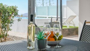 A table with a bottle, a potted plant, a bowl of fruit, and two glasses, set outdoors with a pool and chairs in the background. - Modern Apartment Casa Amanecer 1 with Pool, Terrace & Wi-Fi; Parking Available (Macher)