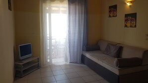 A living room with a sofa, a television on a stand, and a window with curtains. - Glyfada Apartment Ag3g 129 (Corfu)
