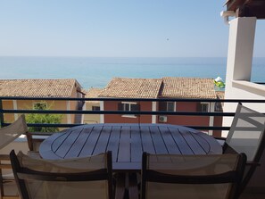 A balcony with a round wooden table and chairs, overlooking a coastal area with buildings and the sea.