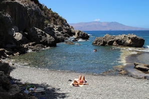 A rocky beach with clear water, a person sunbathing, and mountains in the background.