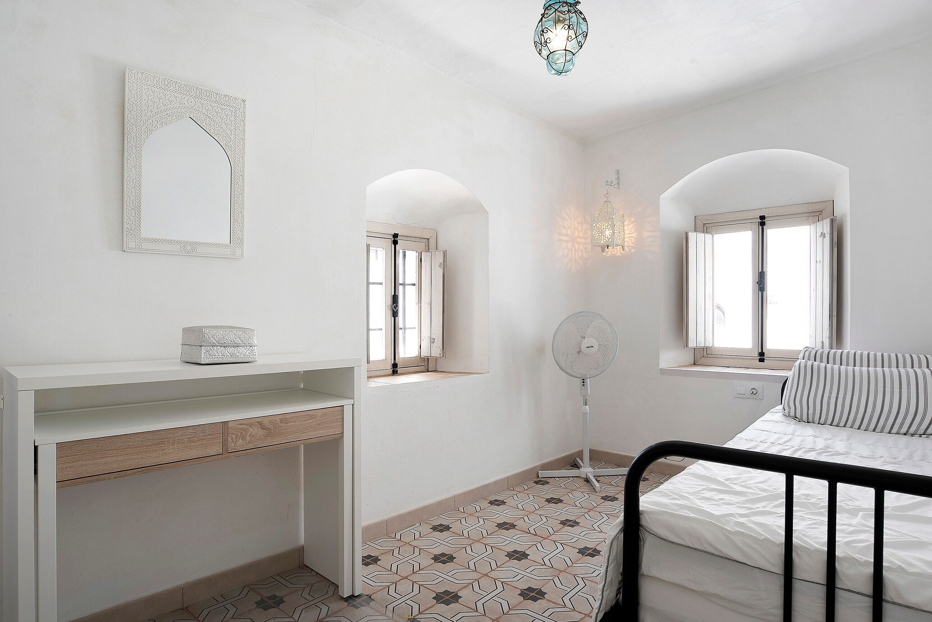 A bedroom with a bed, a bedside table, a desk, a chair, a standing fan, and a decorative mirror.