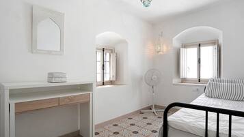 A bedroom with a bed, a bedside table, a desk, a chair, a standing fan, and a decorative mirror.
