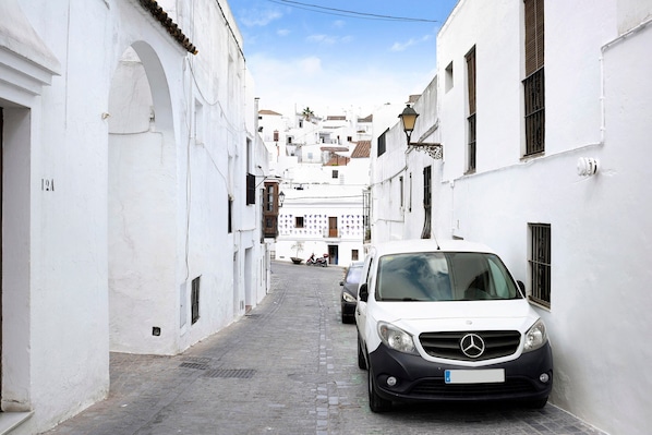 Une rue étroite bordée de bâtiments blancs, une voiture garée et un ciel dégagé.