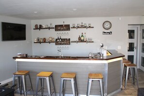 A bar area with a wooden countertop, stainless steel stools, and shelves with bottles and glasses.