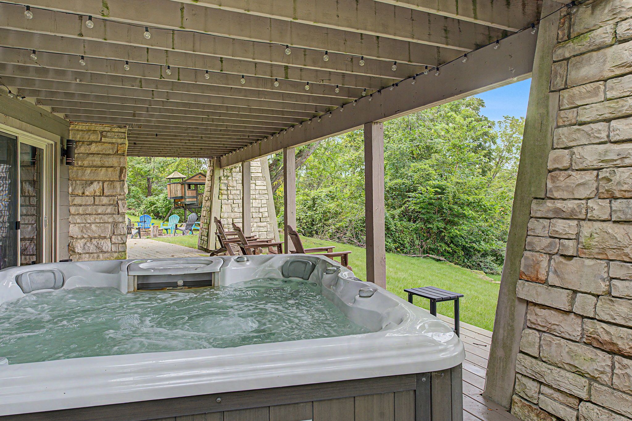 A hot tub under a wooden pergola with stone walls, surrounded by outdoor furniture and a play structure.