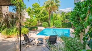 Un área de piscina con sillas y una pequeña mesa, rodeada de exuberante follaje y palmeras.