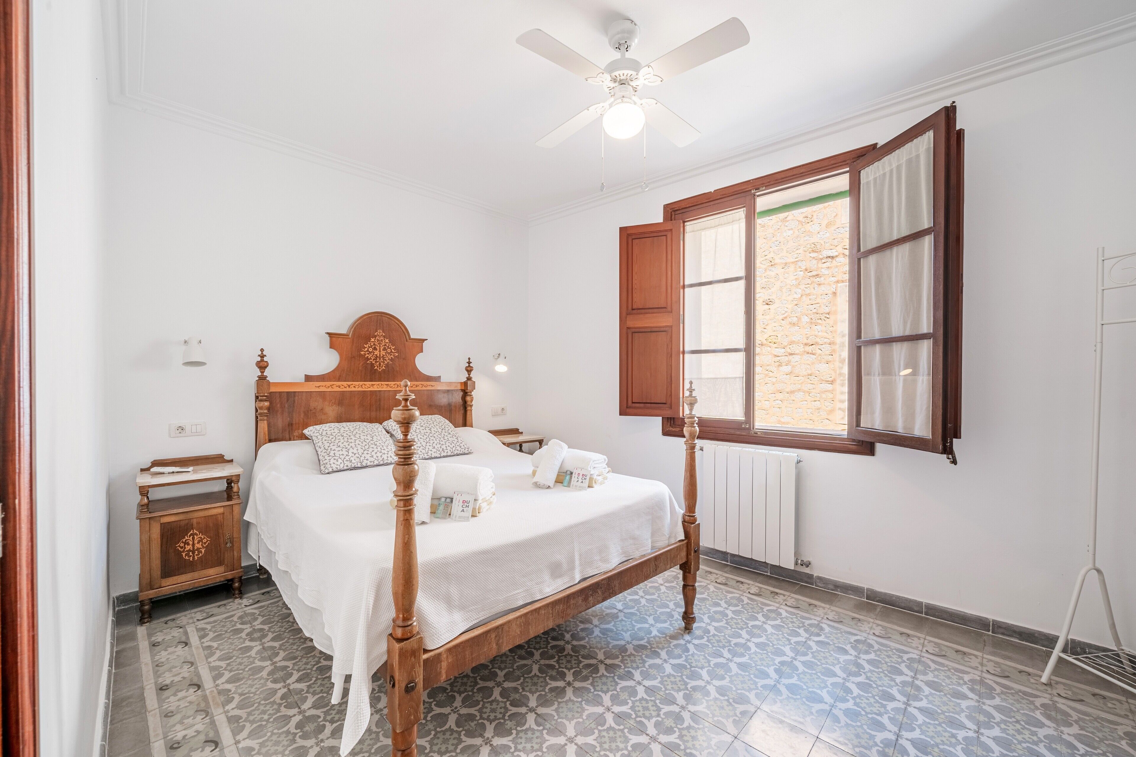 Un dormitorio con cama con dosel, ventilador de techo, ventana con contraventanas de madera y baldosas de suelo estampadas.