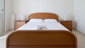 A neatly made bed with white linens and a folded towel, two bedside tables with lamps, and a large window on one side. - Manoleas Villas- Apartment 1 (West Mani)