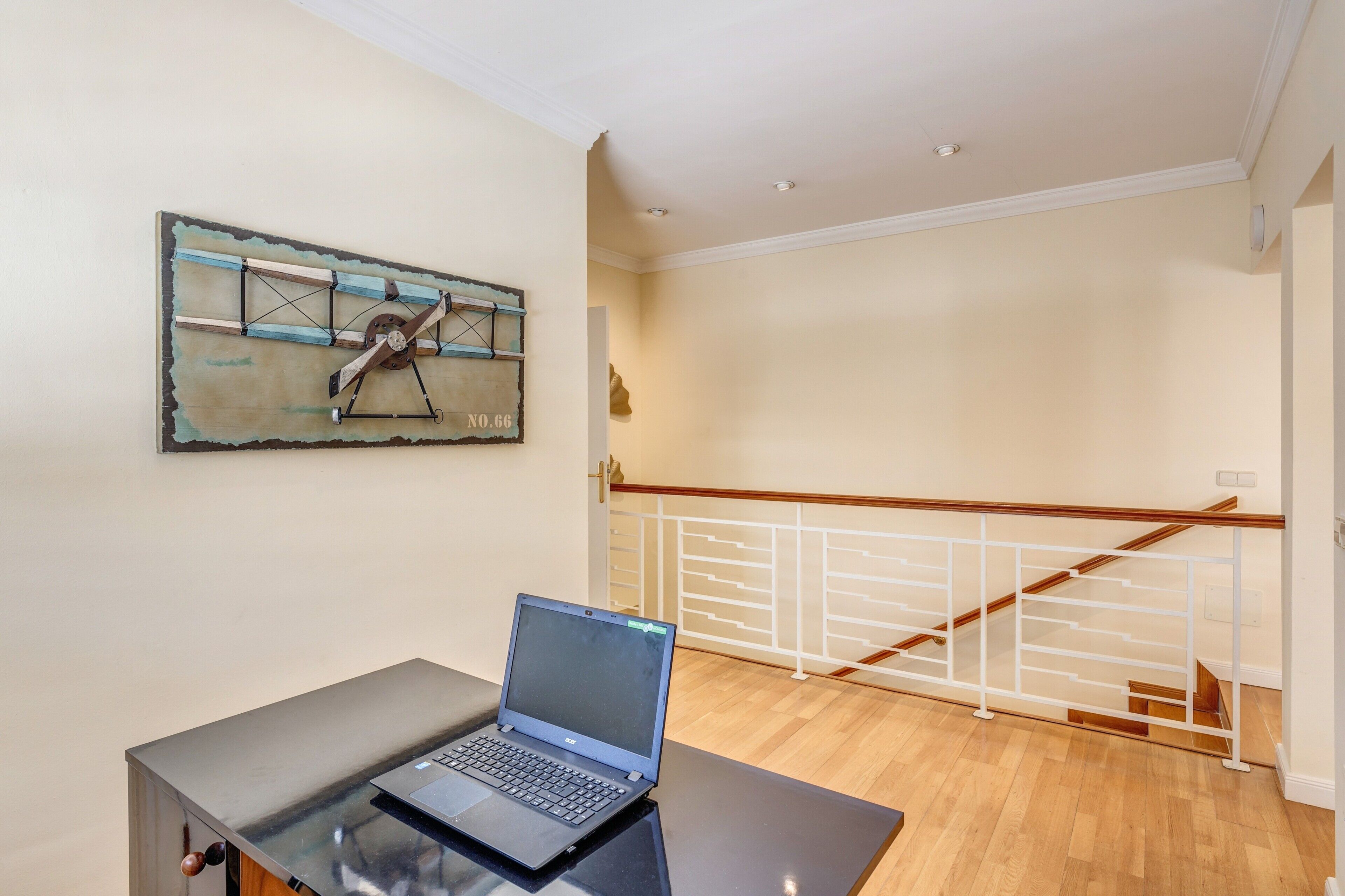 Un portátil sobre una mesa, una pared con una obra de arte rústica y una escalera con barandilla blanca.