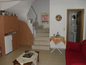 A living room with a red sofa, a small table with a tablecloth, a kitchen area with cabinets and a refrigerator, a staircase, and a bathroom visible through a doorway.