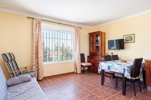 A room with a sofa, dining table, chairs, a cabinet, a television, and a window with curtains.