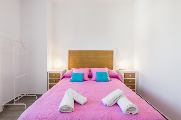 A neatly made bed with pink bedding and two blue pillows, two bedside tables with lamps, and a wooden headboard.