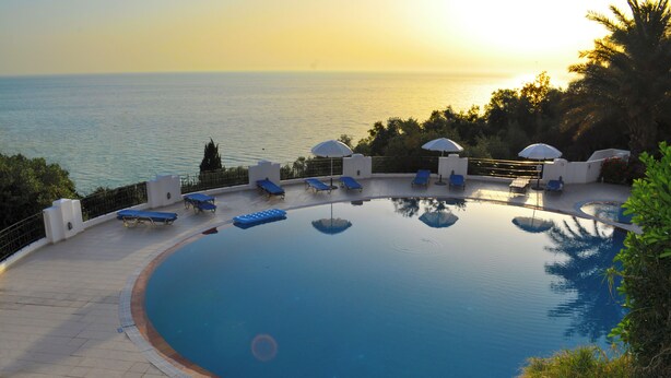 Une piscine à débordement circulaire, dotée de chaises longues et de parasols, offrant une vue sur une étendue d’eau au coucher du soleil.