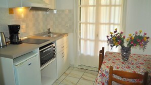 Une petite cuisine équipée d’une machine à café, d’un four et d’un évier. On y trouve une table à manger recouverte d’une nappe à motifs floraux et agrémentée d’un vase de fleurs.