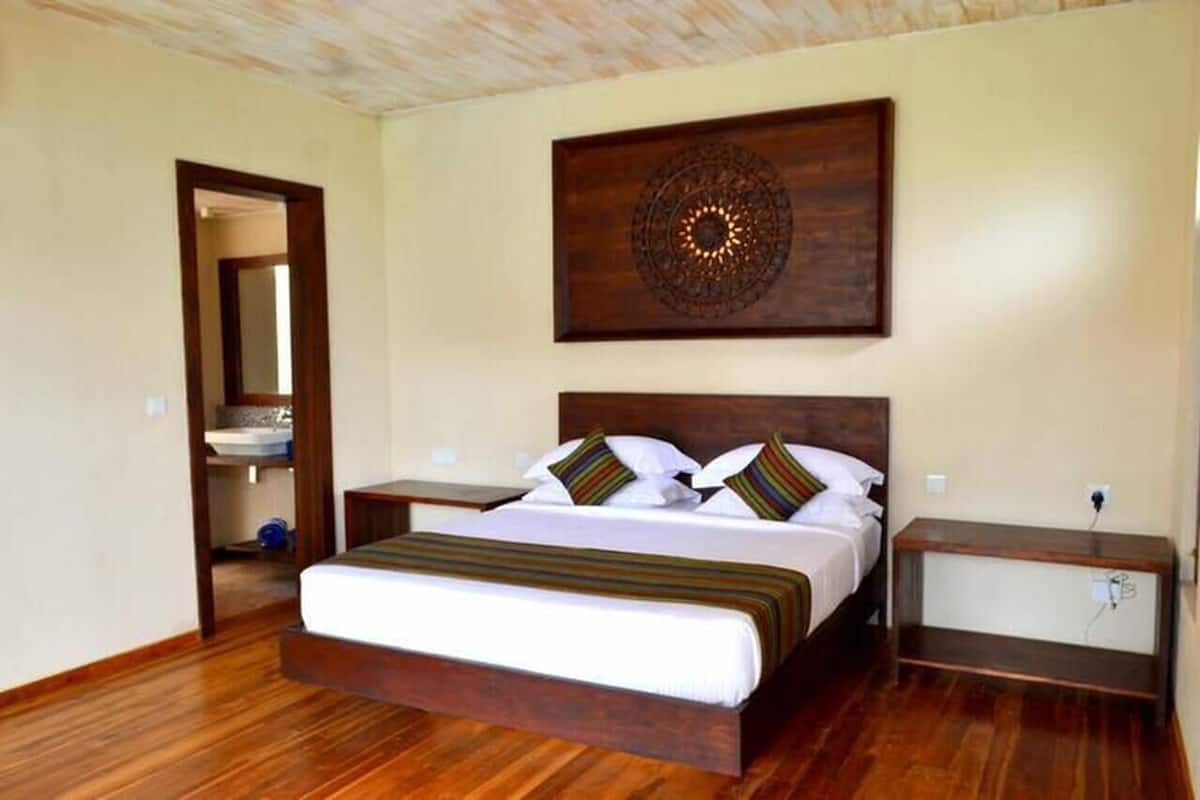 A bedroom with a wooden bed, a bedside table, a nightstand, a wall-mounted decorative piece, and a door leading to a bathroom.