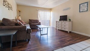A living room with a sofa, armchair, coffee table, TV stand, and a window with blinds.