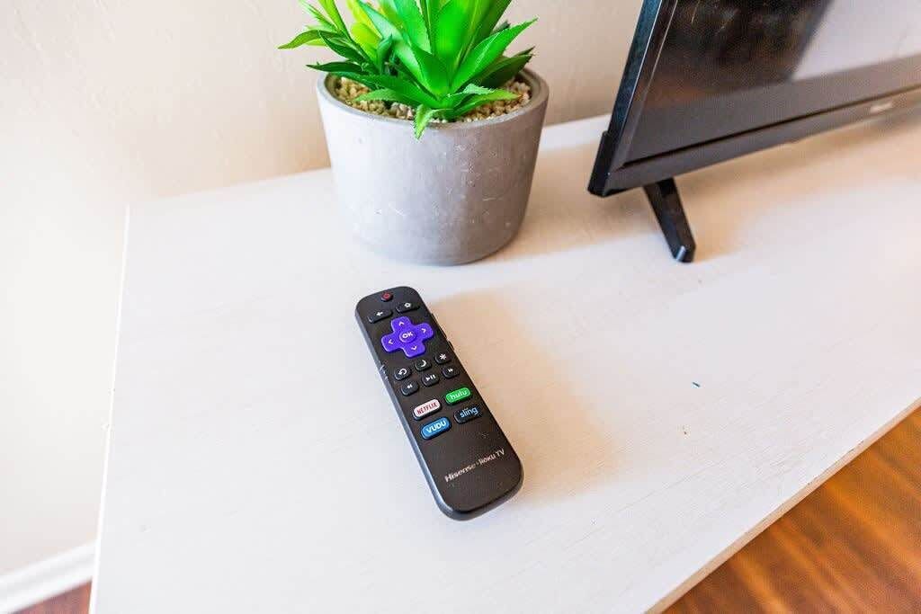 Valkoisella alustalla on kaukosäädin, ja taustalla näkyy ruukkukasvi ja televisio.