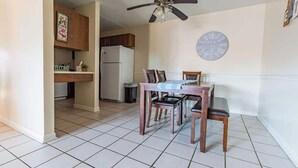 A dining area with a table and chairs, a ceiling fan, a clock, and a kitchen in the background.