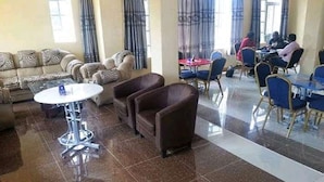A hotel lobby with a seating area, a dining section, and a group of people in the background. - Downhill Springs Resort (Webuye)