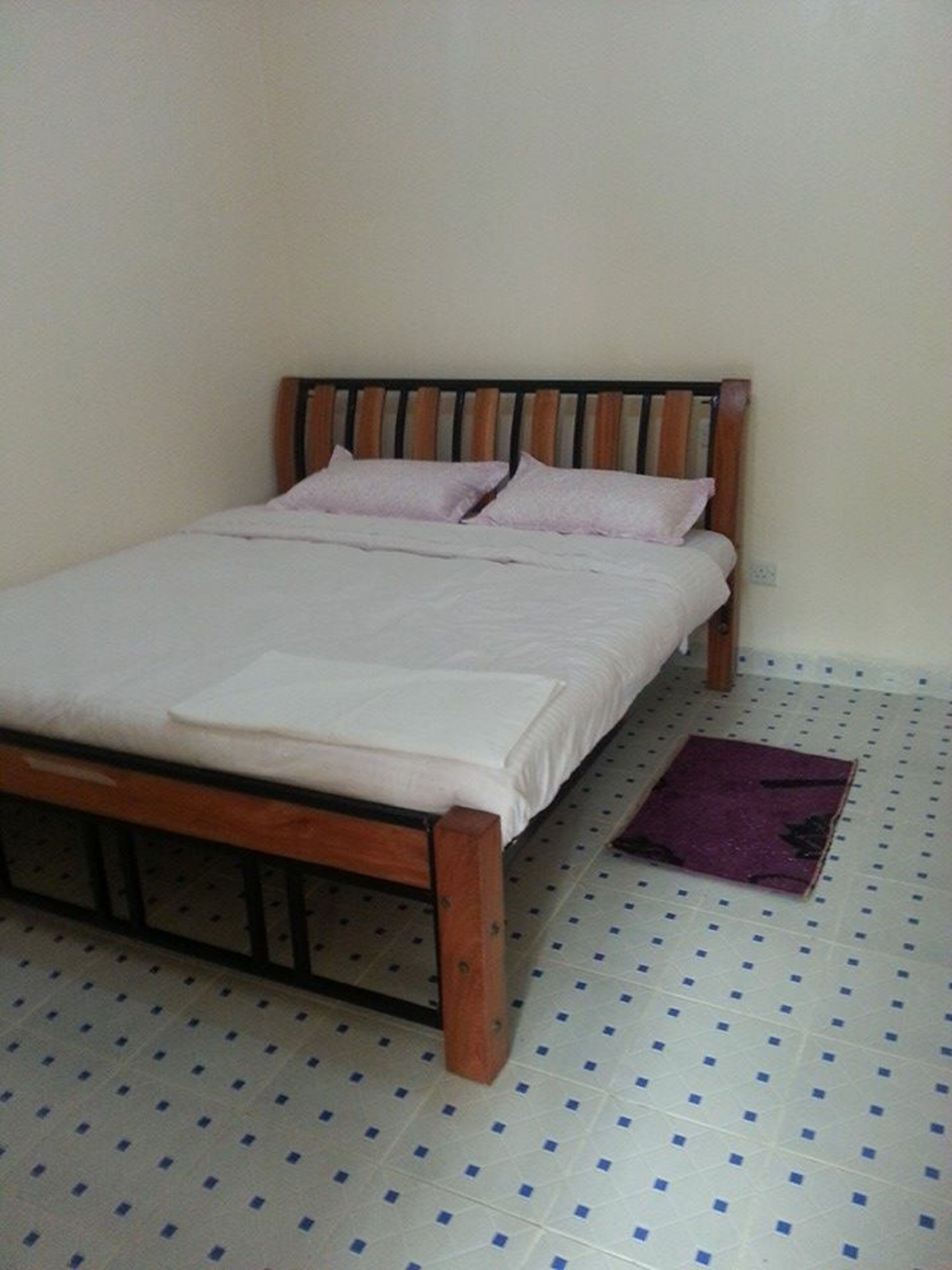 A single bed with a wooden frame and a mattress, a purple rug on the floor, and a white wall.